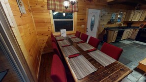 A wooden interior with a dining table set for six, red upholstered chairs, a kitchen area with wooden cabinets, and a window with a view of trees.