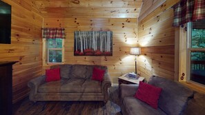 A cozy cabin interior with wooden walls, a fireplace, two sofas with red cushions, and a painting of a forest scene. - Serendipitous Luxury Cabin with Forest Views, Hot Tub, mins from Hocking Hills (Logan)