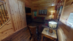 A cozy cabin interior with wooden walls, a sofa, a table with a lamp, and a window with curtains. - Serendipitous Luxury Cabin with Forest Views, Hot Tub, mins from Hocking Hills (Logan)