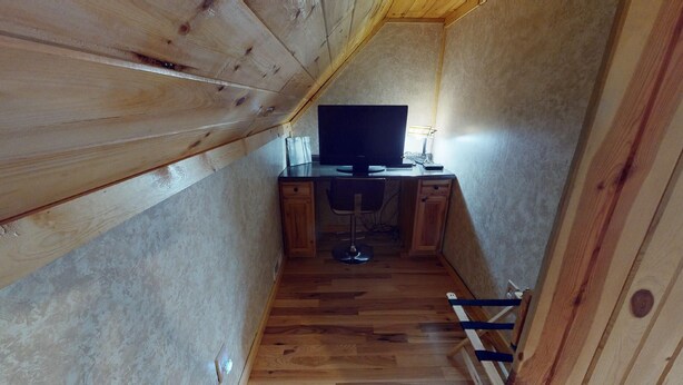 Una habitación estrecha con paredes de madera y techo inclinado, que incluye un escritorio con una computadora y una silla, además de una escalera apoyada en la pared.