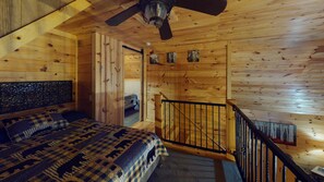 A wooden cabin bedroom with a bed, a ceiling fan, and a staircase.