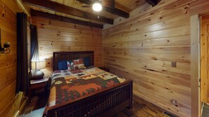 Une chambre dans une cabane en bois, avec un lit, une table de chevet, une lampe et un ventilateur de plafond.