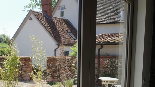 A view through a glass door into a backyard with a white table and chairs, a brick wall, and a house with a tiled roof. - The Cartlodge near Woodbridge - recently converted barn (Woodbridge)