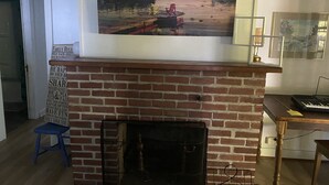 Une pièce avec une cheminée en briques, un tableau représentant un paysage encadré au-dessus, un petit tabouret bleu et un clavier posé sur une table en bois.