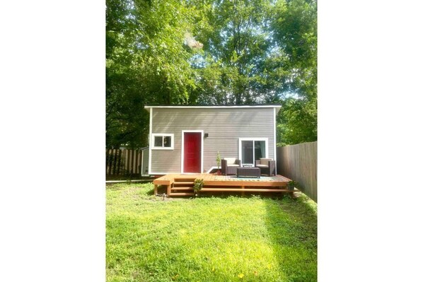 A small, single-story house with a red door, a deck, and a fenced backyard.