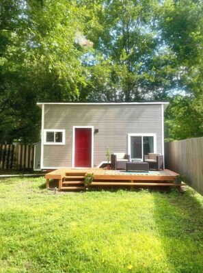 A small, single-story house with a red door, a deck, and a fenced backyard.