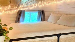 A bed with white bedding, a black metal frame, and a white rug. There is a window with curtains, and string lights are visible on the ceiling. - 🏡Cozy Farmhouse Tiny Home in Backyard Pool Oasis🌻 (Austin)