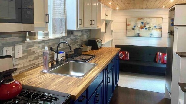 A modern kitchen with a wooden countertop, a sink, and a stove. There is a window with a curtain, a painting on the wall, and a bunk bed in the background. - 🏡Cozy Farmhouse Tiny Home in Backyard Pool Oasis🌻 (Austin)