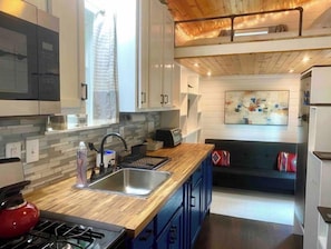 A modern kitchen with a wooden countertop, a sink, and a stove. There is a window with a curtain, a painting on the wall, and a bunk bed in the background.
