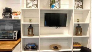 A modern living room with a flat-screen TV mounted on a white wall, surrounded by shelves with decorative items and books. - 🏡Cozy Farmhouse Tiny Home in Backyard Pool Oasis🌻 (Austin)
