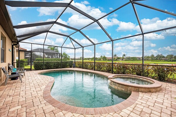 A covered swimming pool with a brick patio, a lounge area with chairs, and a view of a residential area.