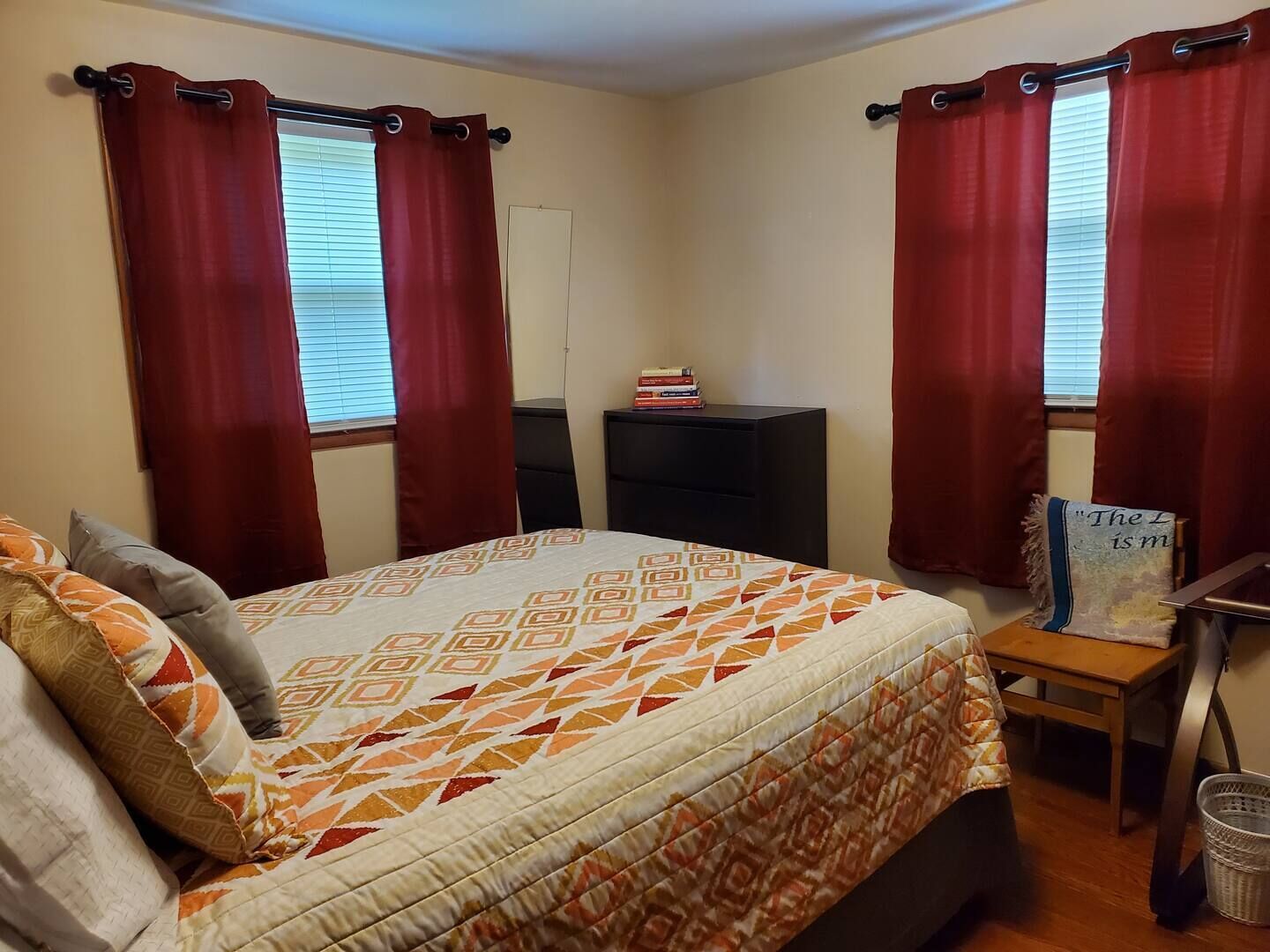 Quarto com cama, duas janelas com cortinas vermelhas, mesa de cabeceira com um livro e uma cadeira pequena.