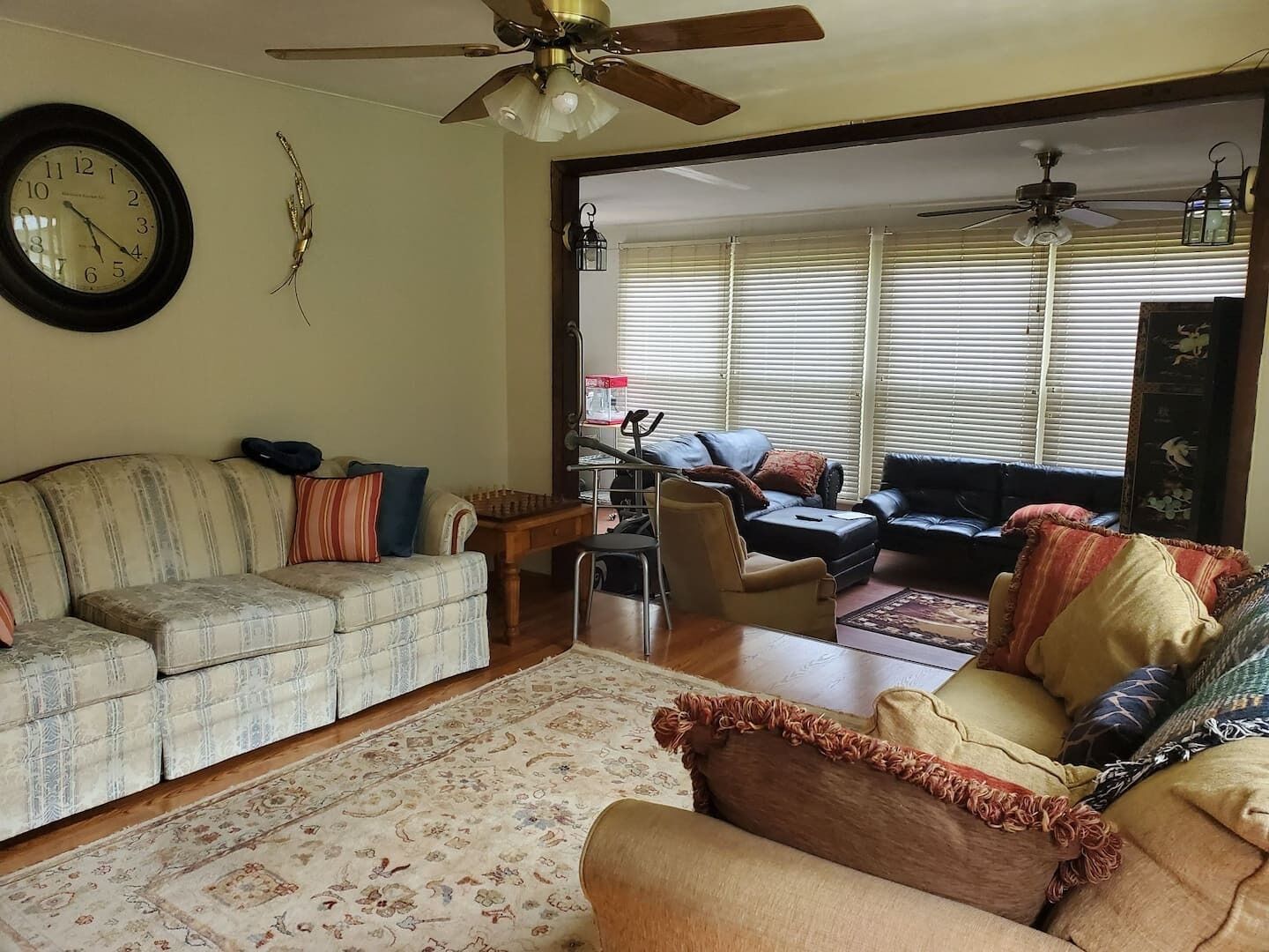 A living room with a sofa, armchairs, a coffee table, and a rug. There is a ceiling fan, a clock, and a window with blinds.