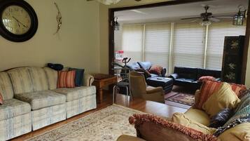 A living room with a sofa, armchairs, a coffee table, and a rug. There is a ceiling fan, a clock, and a window with blinds.