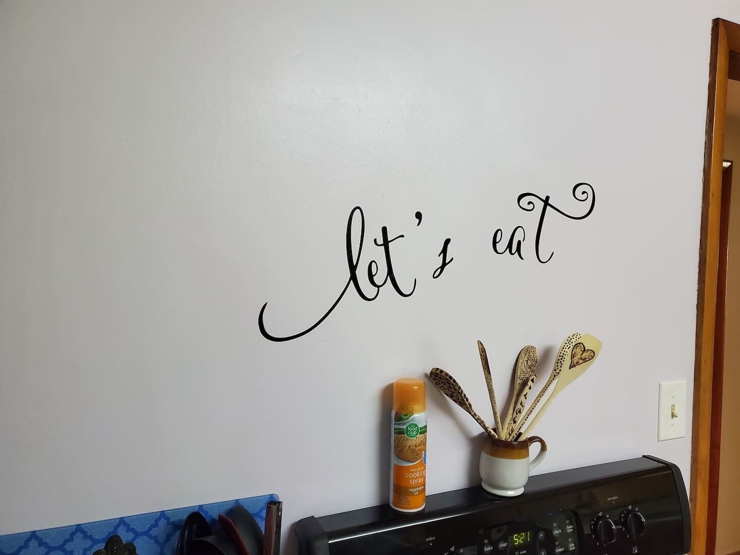 Pared de cocina con un cartel que dice 'comamos', una lata de aerosol de cocina y una taza con utensilios.