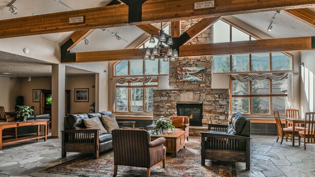 A spacious living room with a stone fireplace, wooden beams, and large windows offering views of the outdoors. - Gateway Lodge 5037 (Keystone)