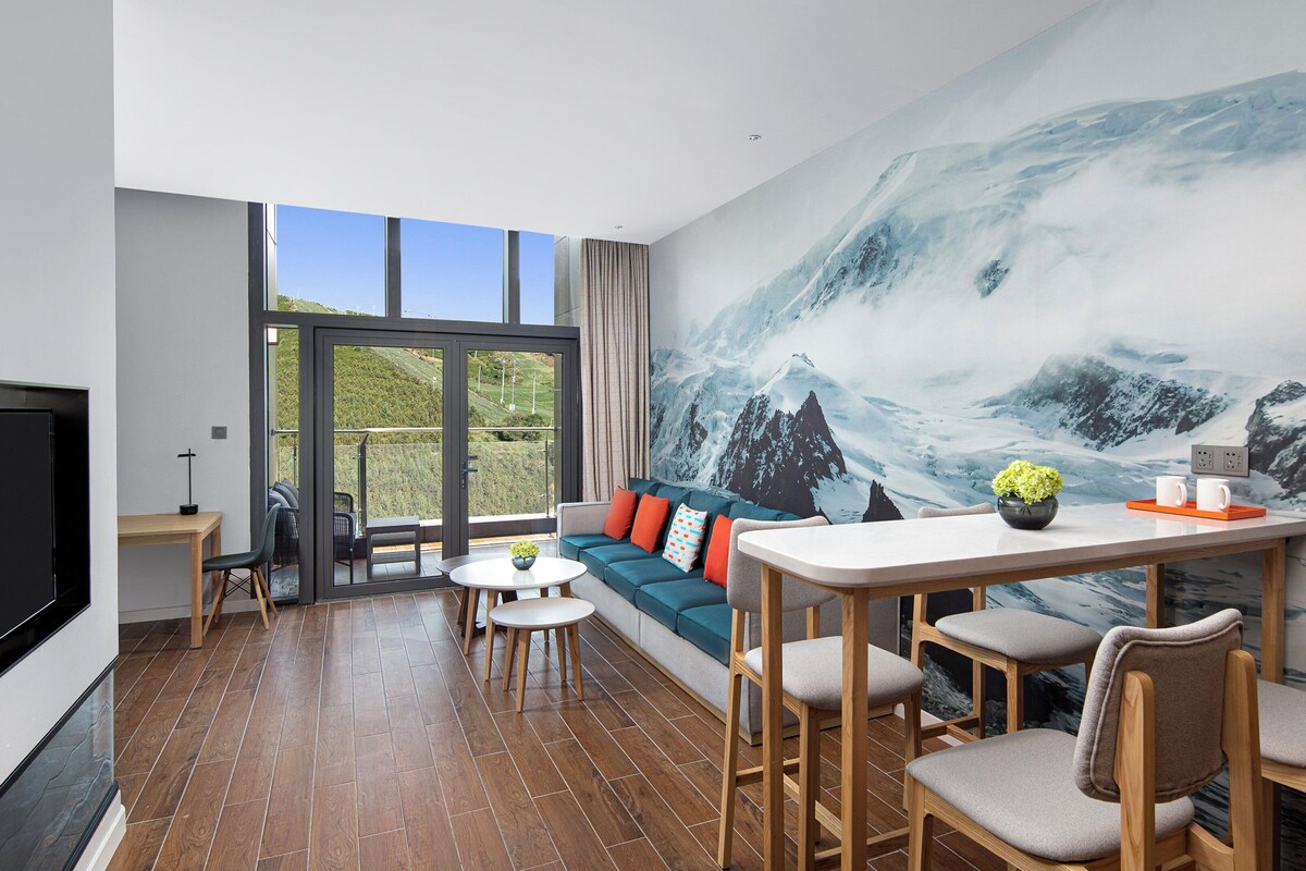 Una habitación de hotel moderna con un ventanal grande, una zona de estar acogedora, una mesa de comedor y un mural con un paisaje de montaña.