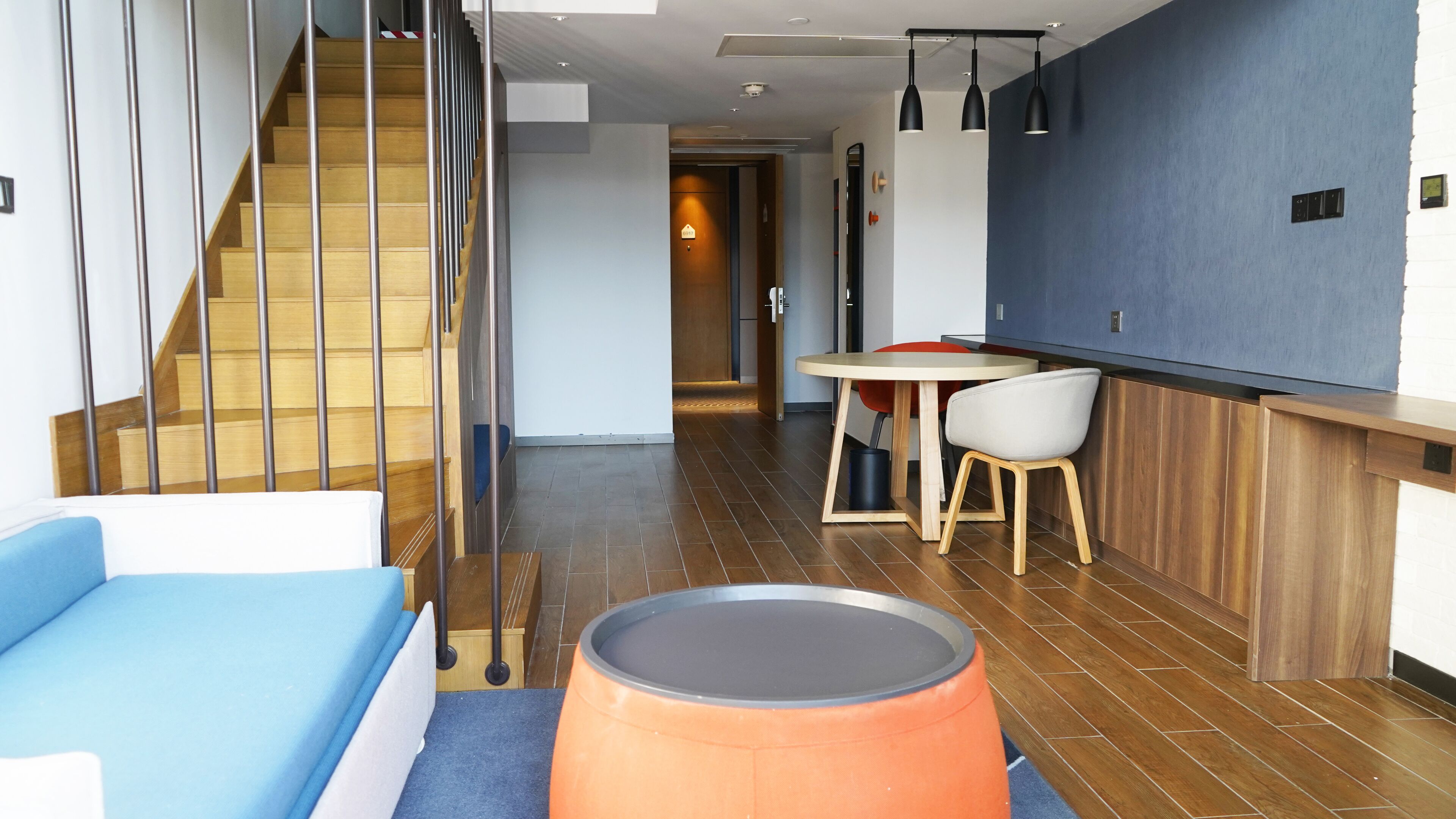 A modern hotel room with a wooden staircase, a blue sofa, a round orange ottoman, and a dining area with a table and chairs.