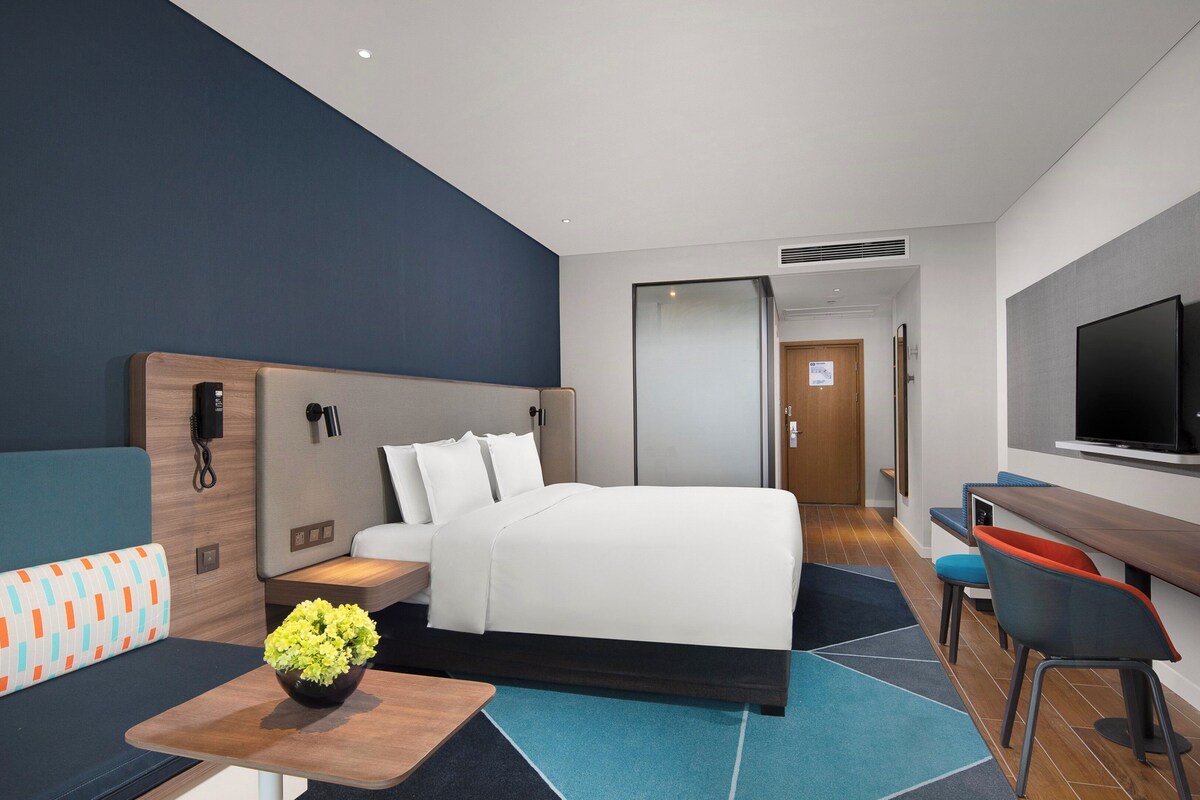 Habitación de hotel moderna con una cama grande, un escritorio, un televisor y un baño visible a través de una puerta abierta.