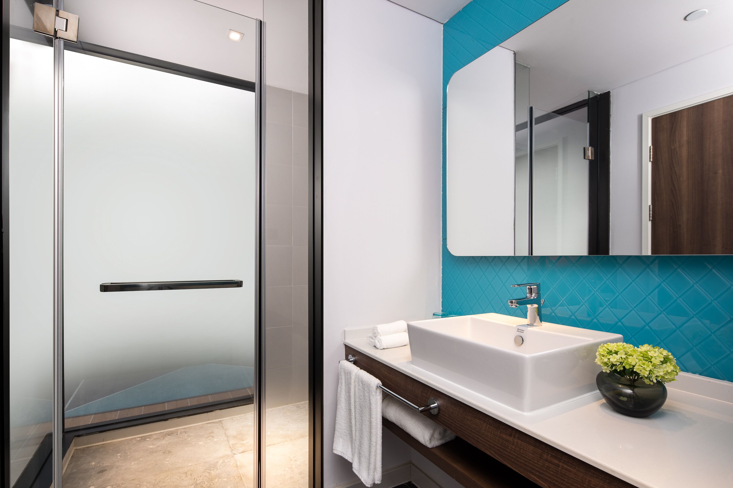 A modern bathroom with a glass shower door, a white sink with a mirror above it, and a teal accent wall.