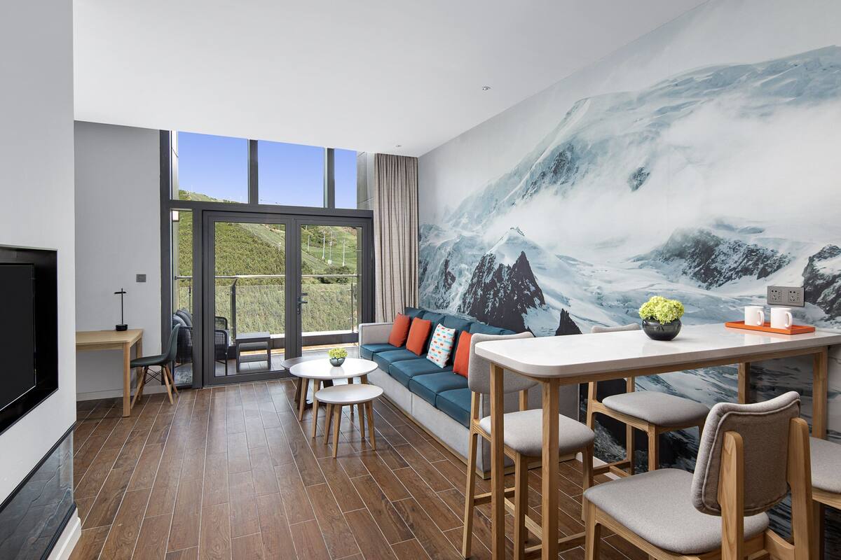 A modern hotel room with a large window, a cozy seating area, a dining table, and a mural of a mountain scene.