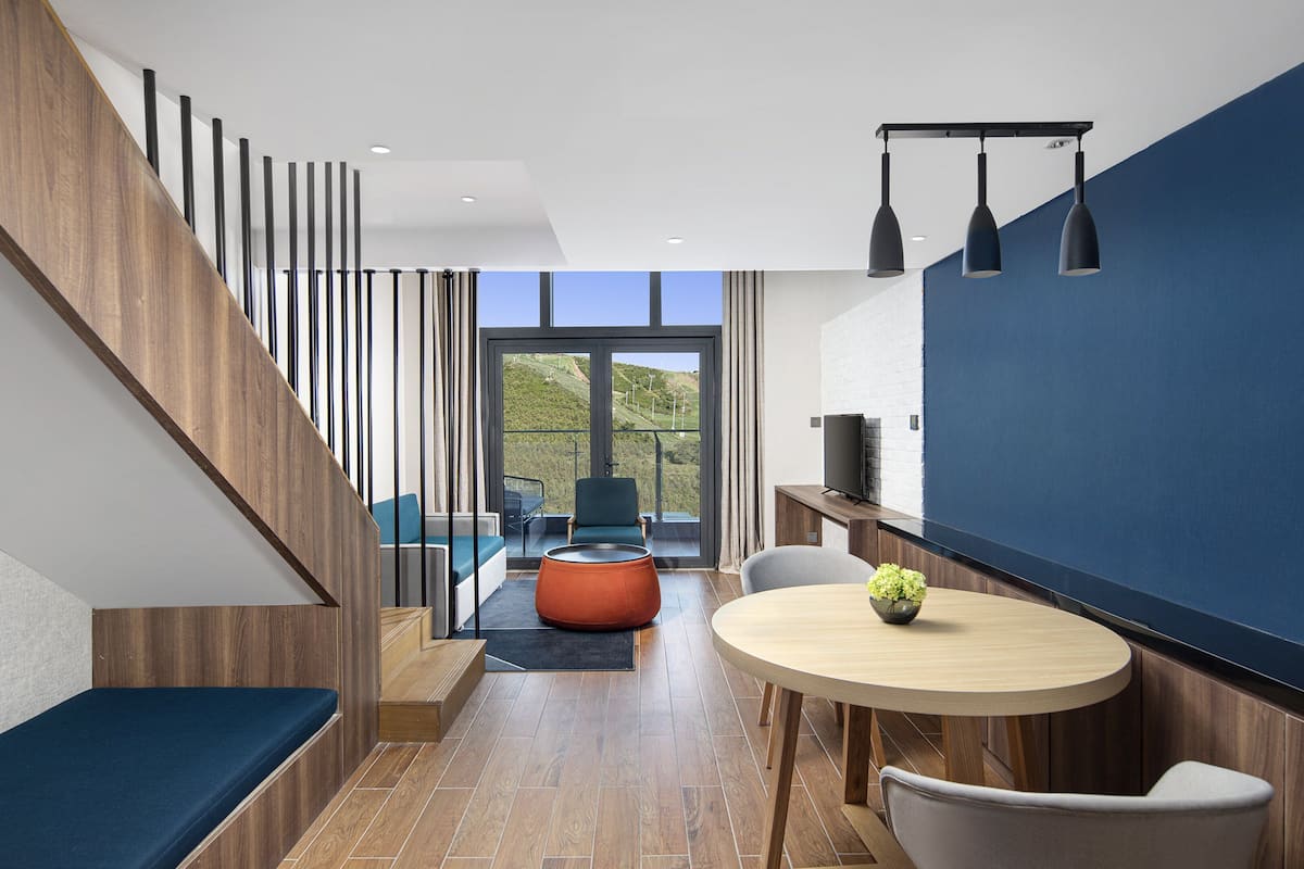 A modern interior with a staircase, a dining area with a round table, and a view of a green landscape through large windows.