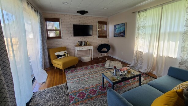 A living room with a yellow armchair, a blue sofa, a patterned rug, a small white table, and a TV mounted on a brick wall.
