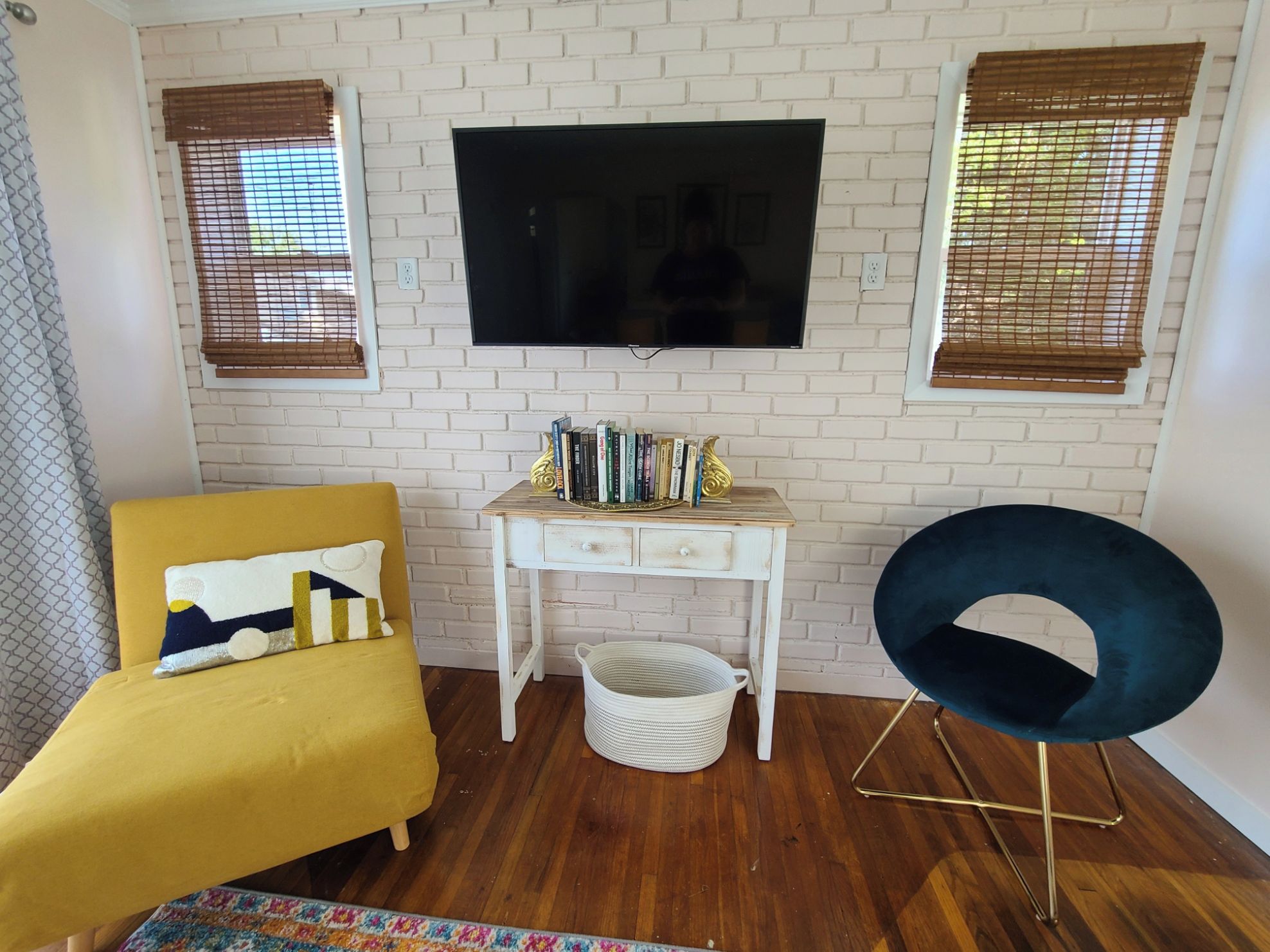 Ein Wohnzimmer mit einem gelben Sofa, einem blauen Sessel, einem weißen Beistelltisch mit Büchern und einem Fernseher an einer weißen Ziegelwand.