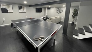 Taula de ping-pong amb un sofà i una televisió a una habitació de parets grises i una escala.