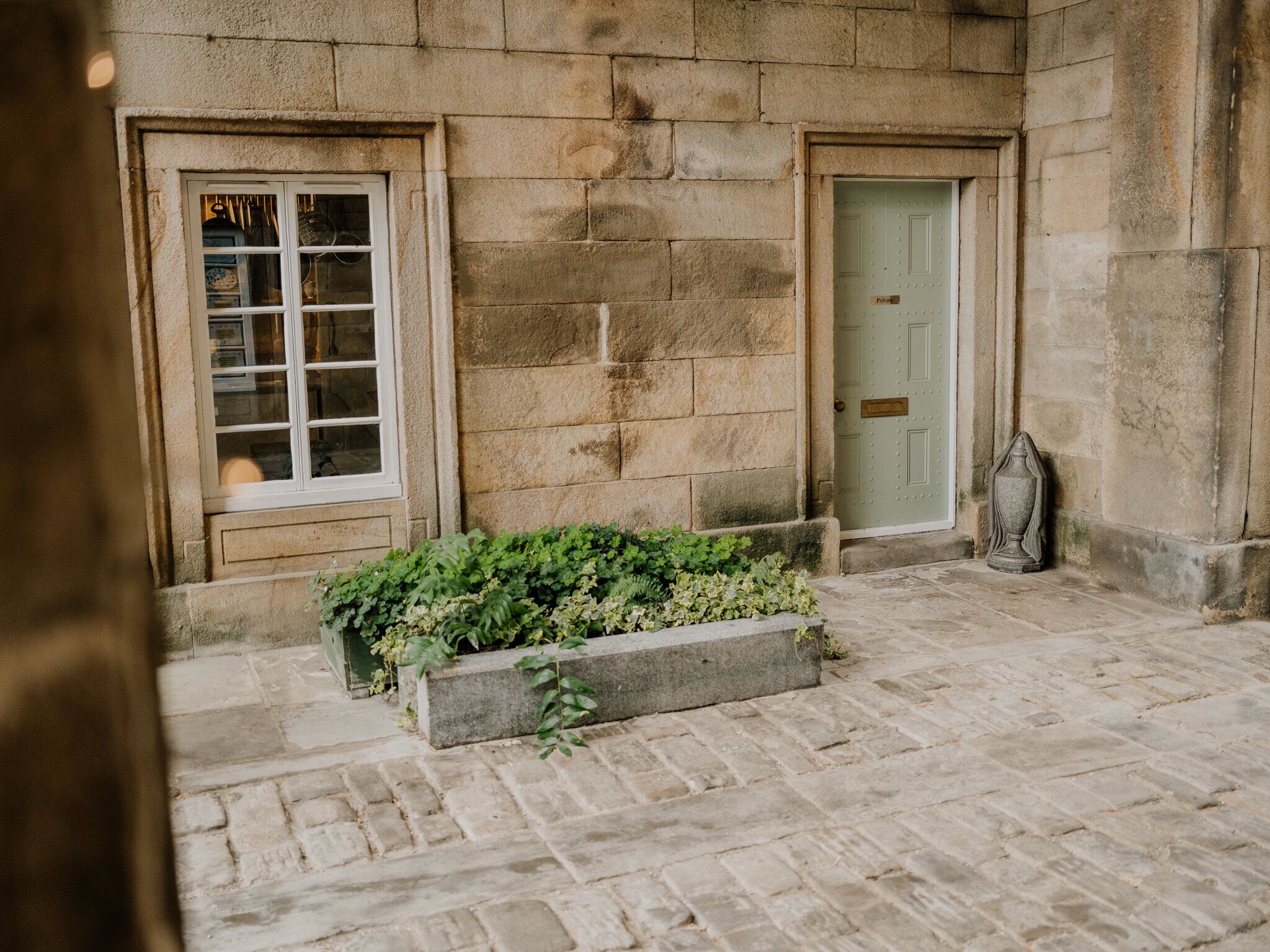 A stone building with two doors and a window, a stone pathway, and a planter with greenery.