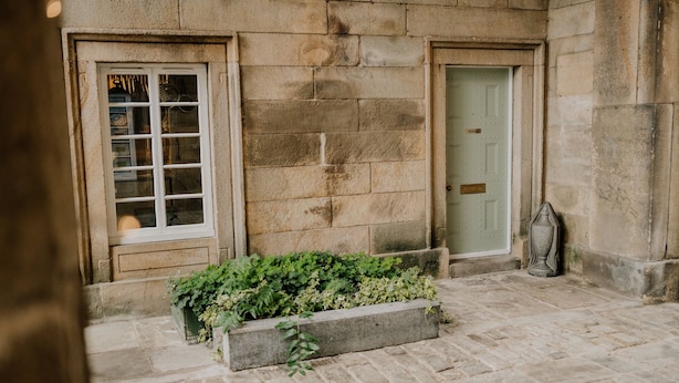 Un bâtiment en pierre doté de deux portes et d’une fenêtre, un chemin en pierre et un bac à fleurs orné de plantes vertes.