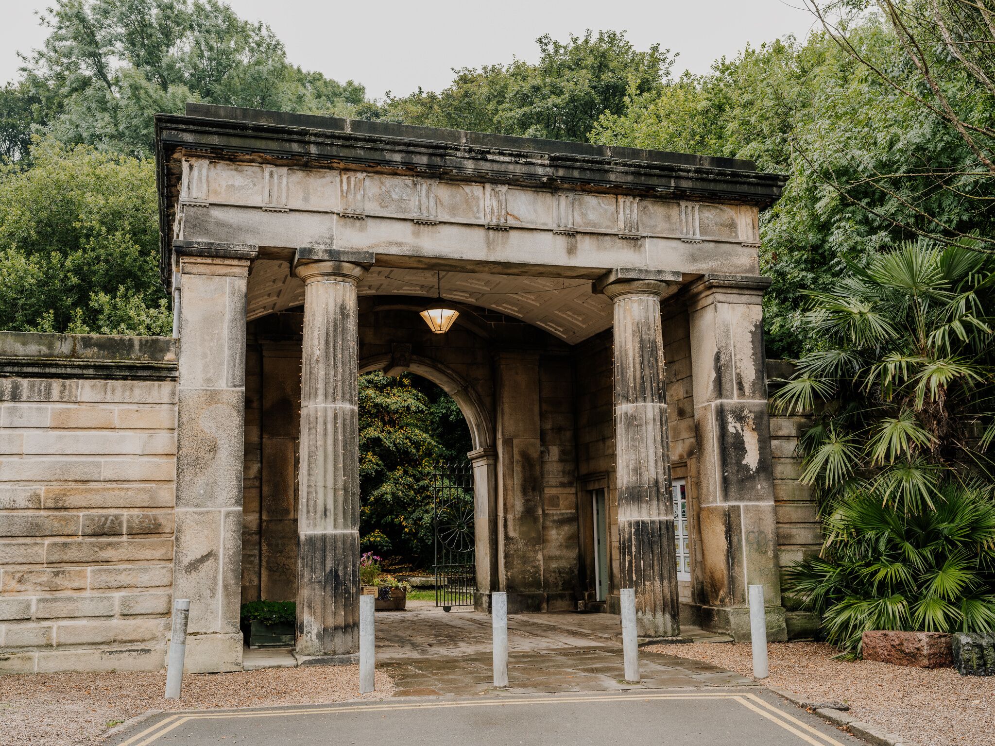 A stone archway with columns, surrounded by greenery and a paved entrance.