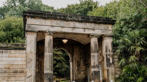 Une arche en pierre, soutenue par des colonnes, entourée de verdure et surmontée d’une entrée pavée.