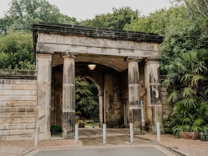Un arco de piedra con columnas, rodeado de follaje y un acceso empedrado.