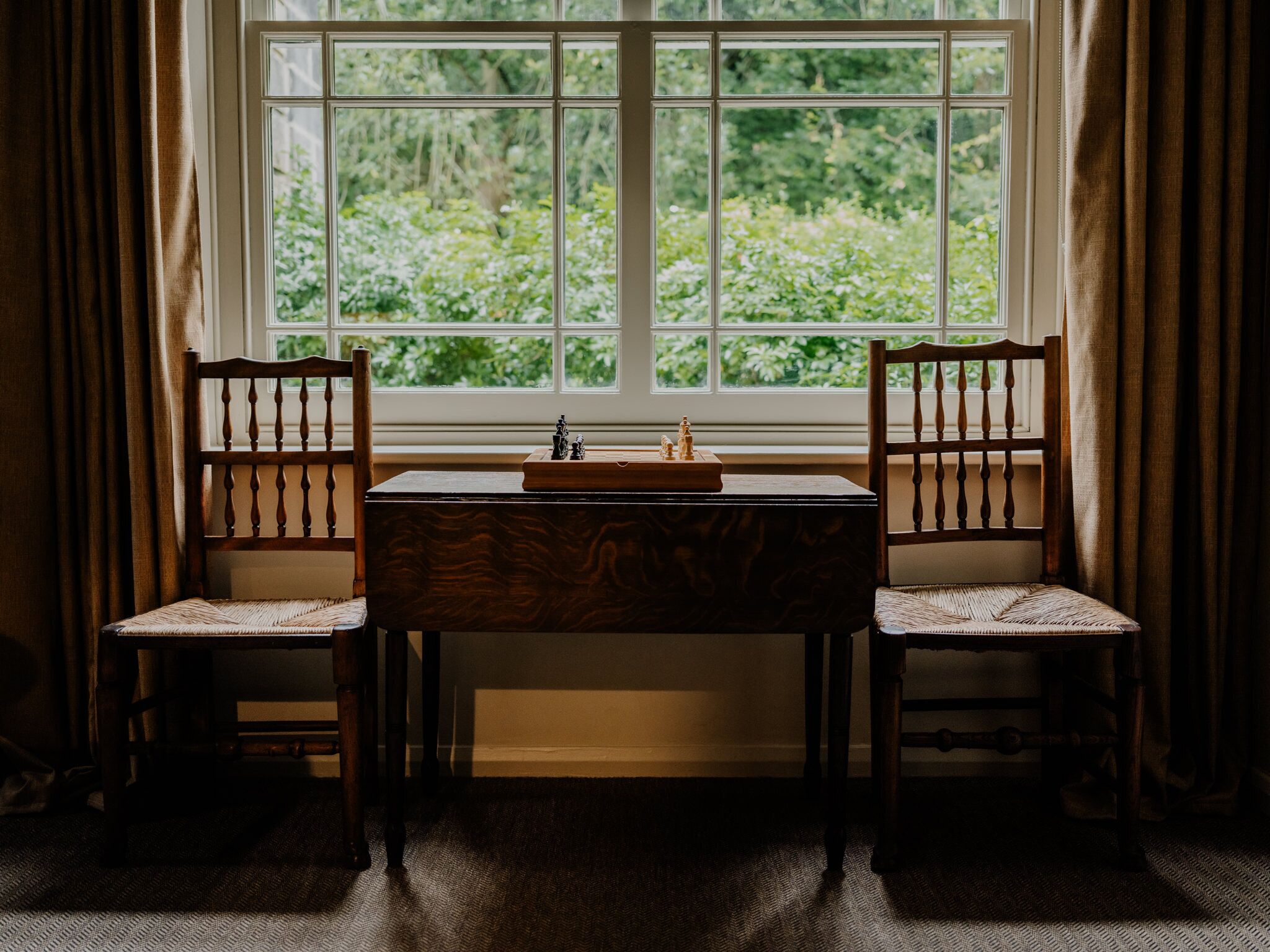A room with a window, two wooden chairs, a table, and a chessboard.