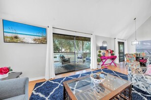 A spacious living room with a glass door leading to a balcony, a dining area with a pink table, and a large beach view painting. - Golf Course 26C - Directly on Golf Course, Including 50% off Golf Daily! (Miramar Beach)