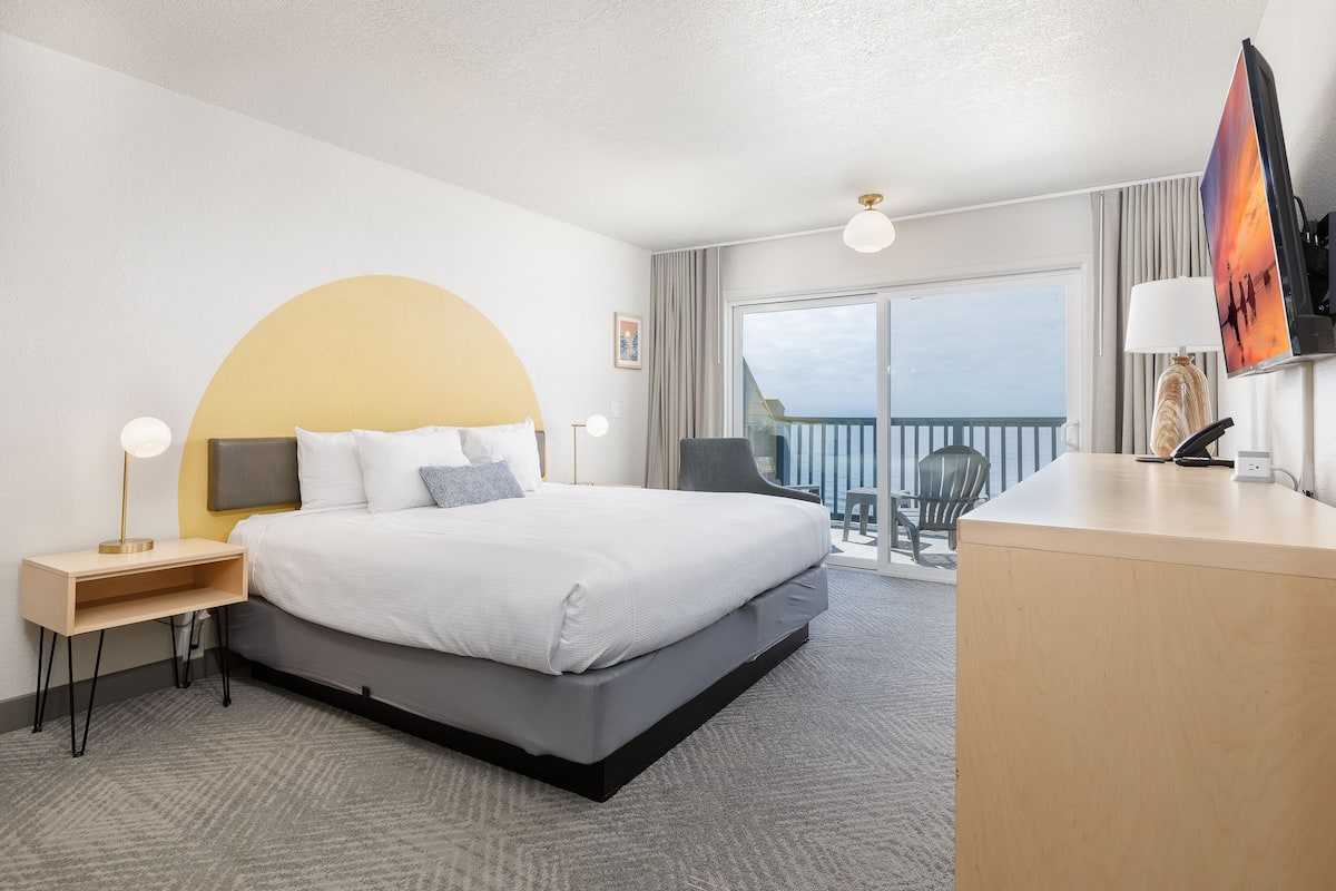 A modern hotel room with a large bed, a desk, a chair, a TV, and a balcony with a view.