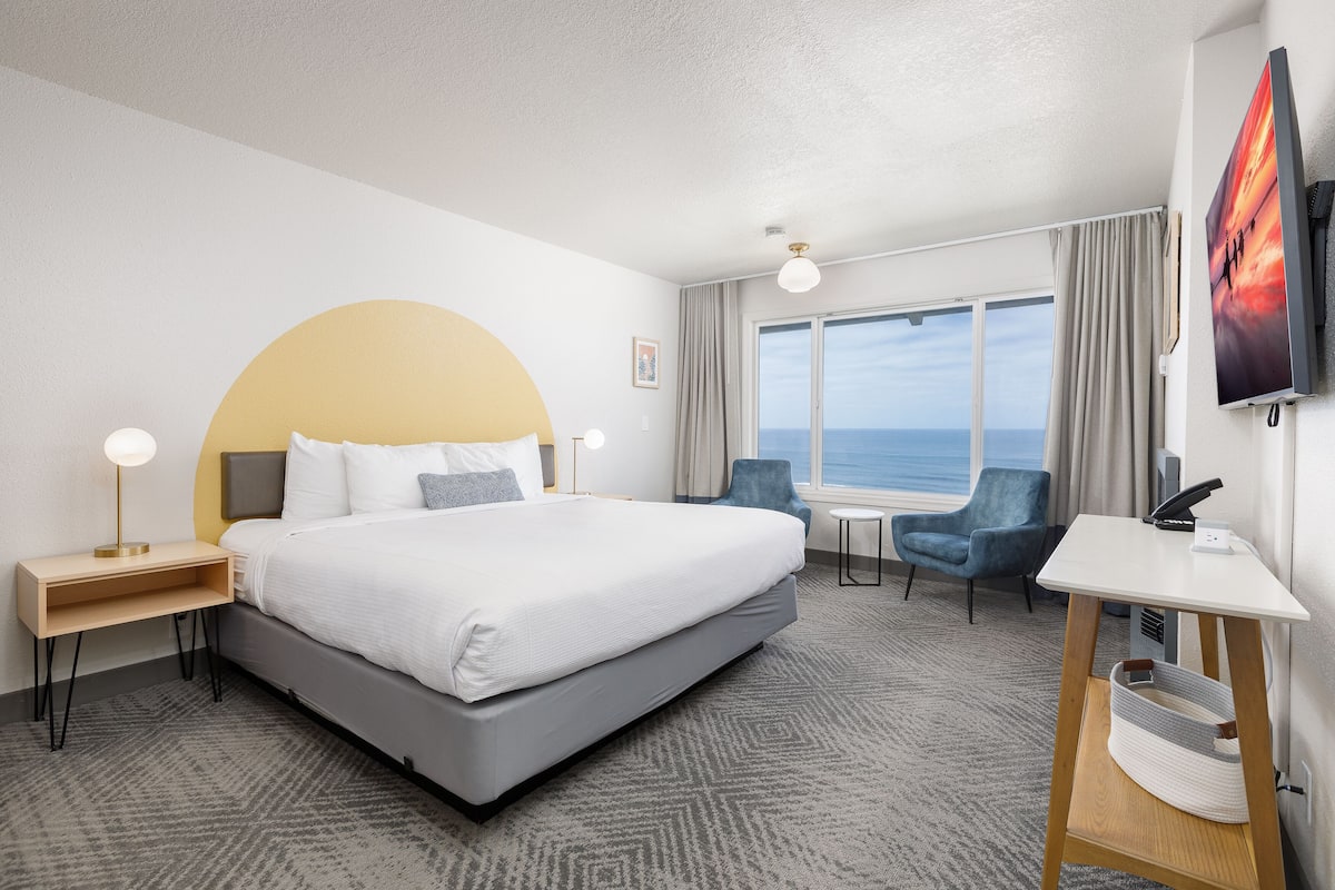 A hotel room with a large bed, a desk, a chair, a TV, and a view of the sea.