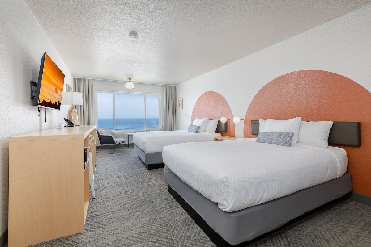 A hotel room with two beds, a desk, a TV, and a view of the ocean.