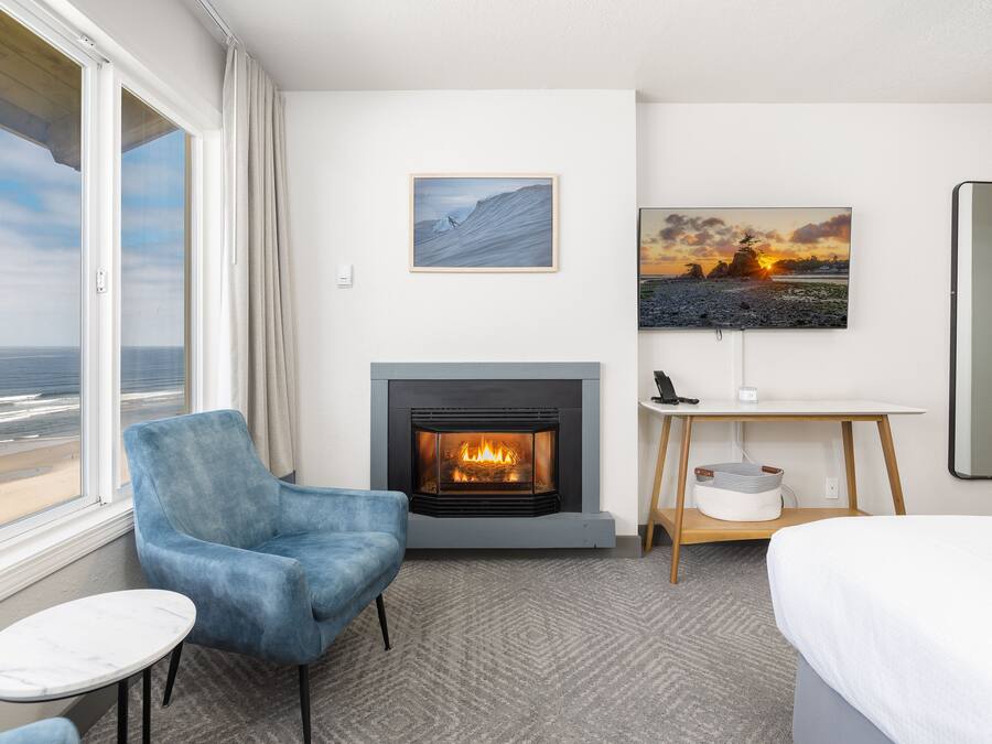 Ett hotellrum med ett stort fönster, en öppen spis, en blå fåtölj, ett litet bord och en säng.