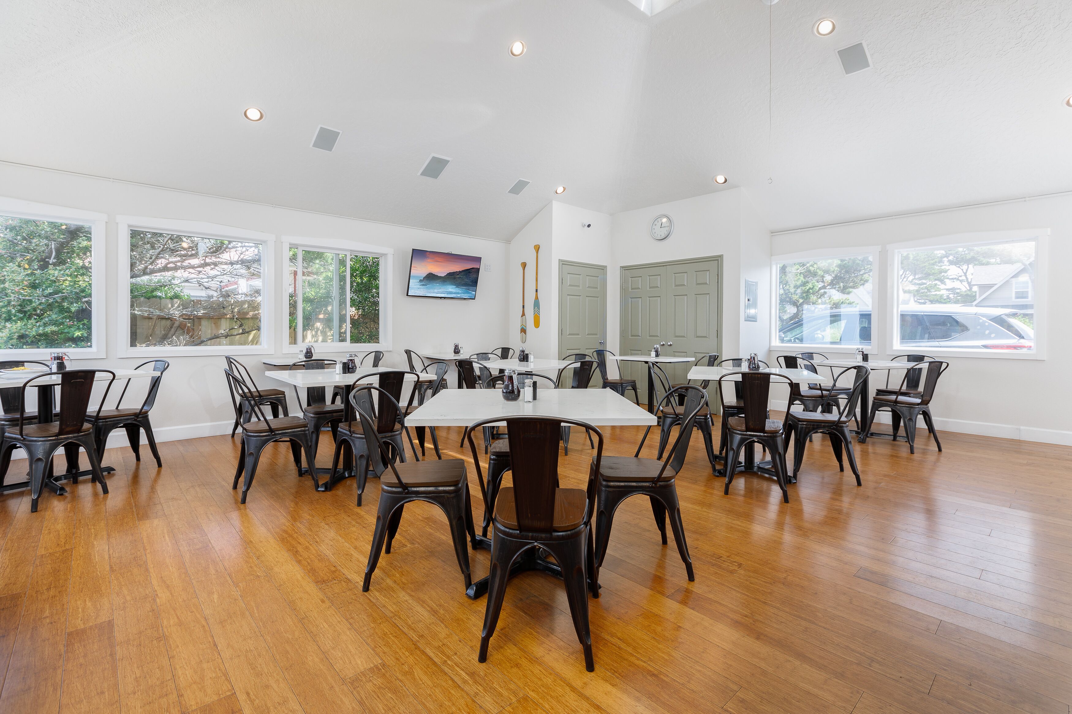 A spacious dining area with multiple tables and chairs, large windows with views of the outside, and a clock on the wall.