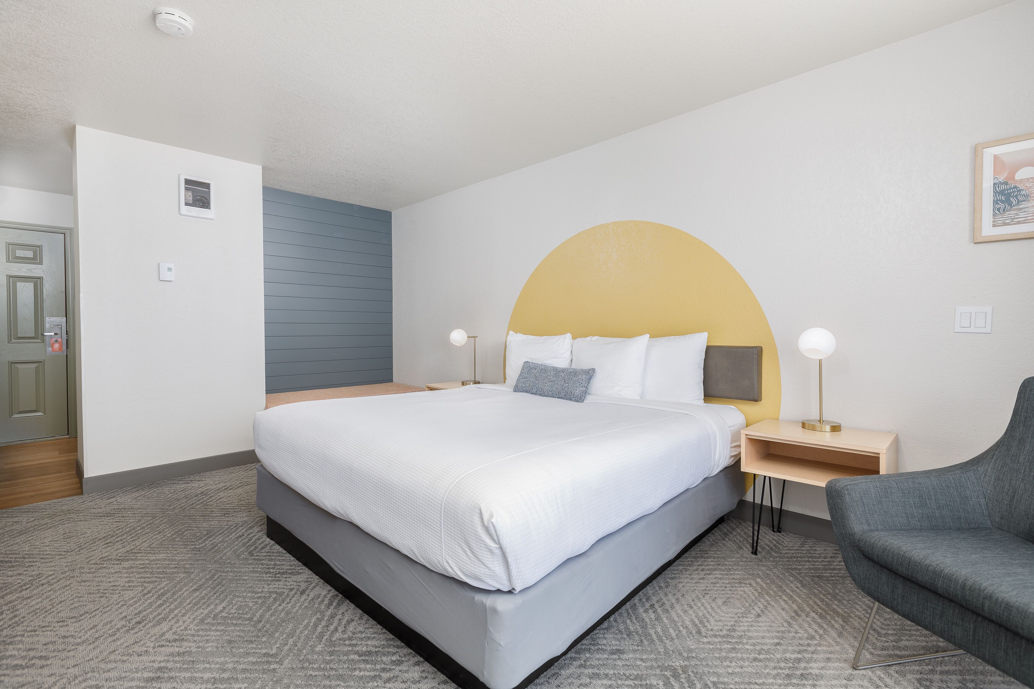 A hotel room with a large bed, a gray armchair, a bedside table with a lamp, and a door with a lock.