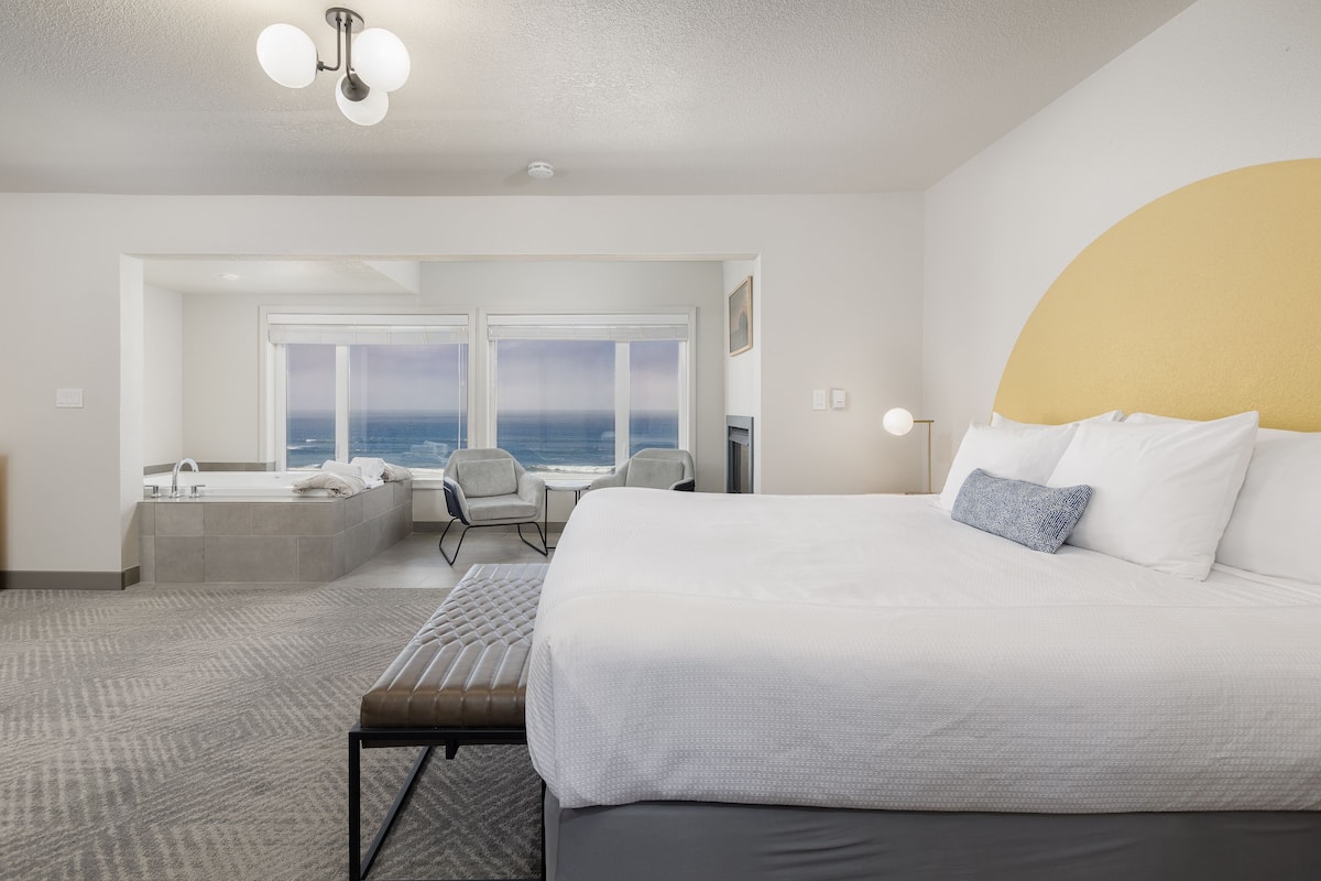 A hotel room with a large bed, a seating area with a view of the ocean, a bathtub, and a desk with a chair.
