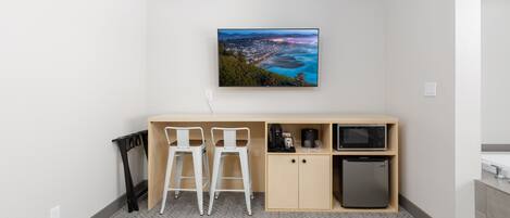 A compact room with a kitchenette, two white bar stools, a small refrigerator, and a wall-mounted flat-screen TV displaying a scenic view.