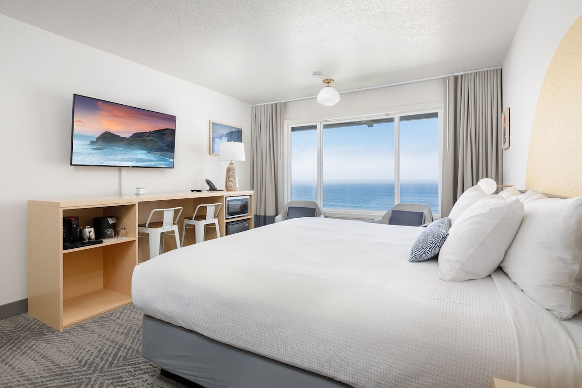 A hotel room with a large bed, a desk with a computer, a TV, and a view of the ocean.