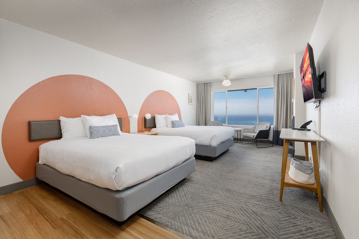 A hotel room with two beds, a desk, a TV, and a large window with a view.