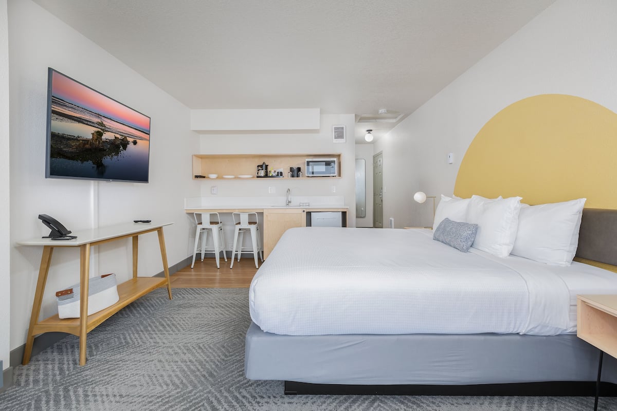A modern hotel room with a large bed, a desk with two chairs, a kitchenette, and a TV mounted on the wall.