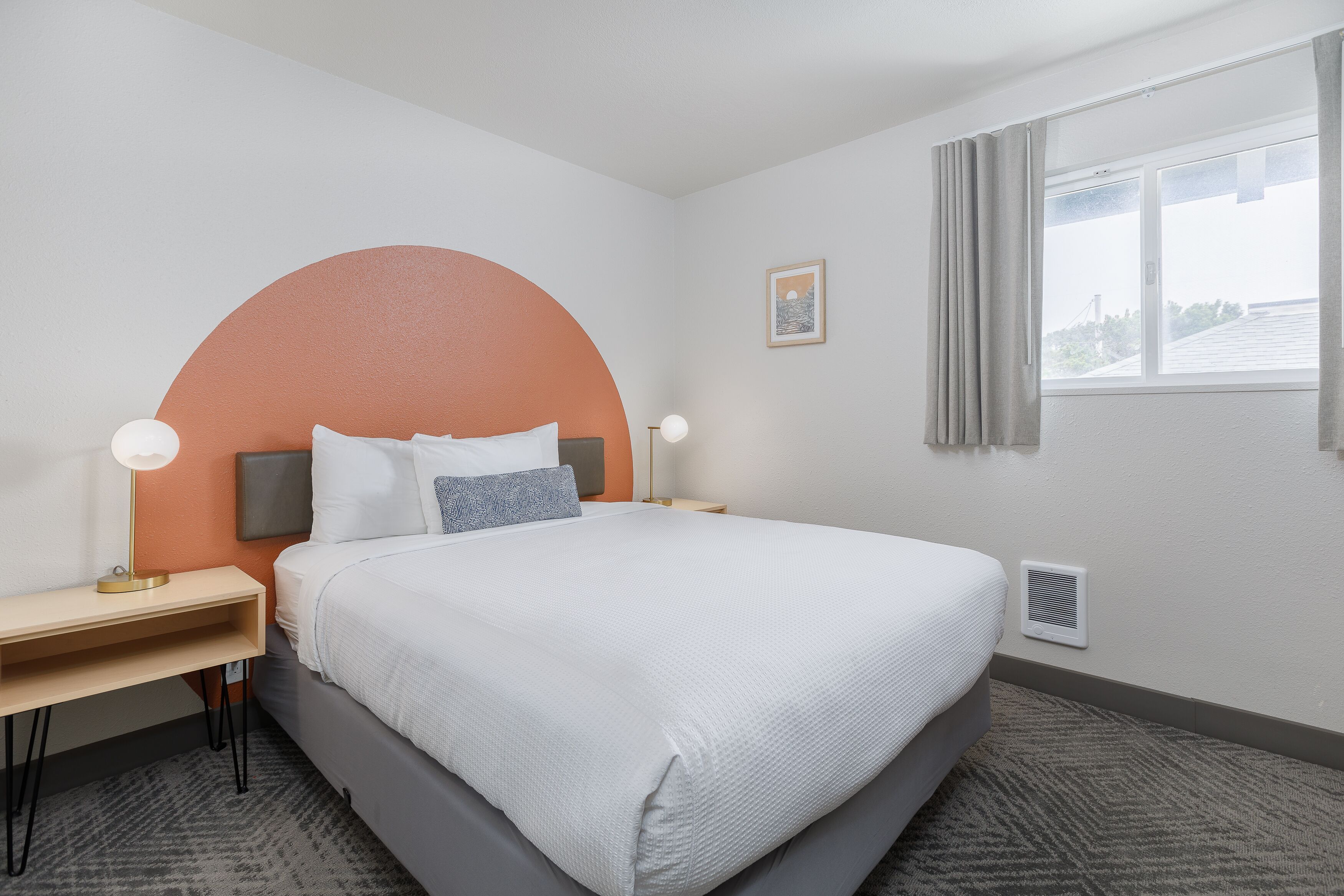 Une chambre d’hôtel avec un lit, un bureau, une lampe, un tableau au mur et une fenêtre avec des rideaux.