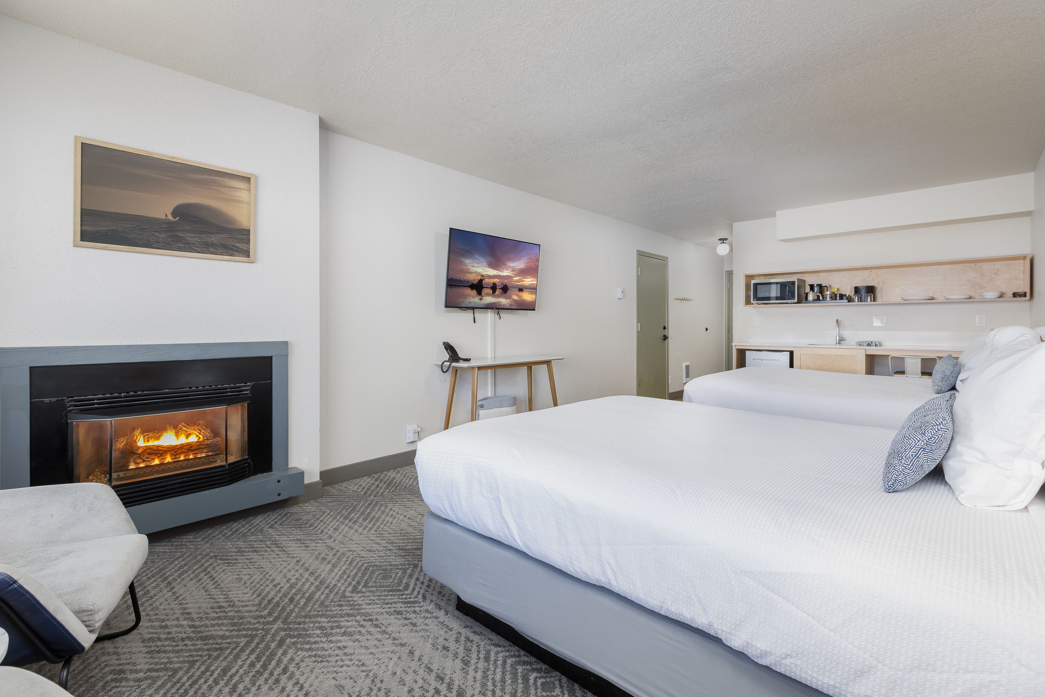 A hotel room with a fireplace, a bed with white bedding, a desk with a lamp, and a painting on the wall.
