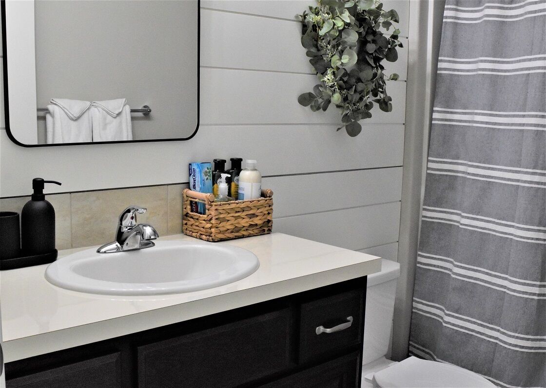 Een badkamer met een witte wastafel, een zwarte kraan en een spiegel erboven. Op het aanrecht staat een rieten mand met toiletartikelen, en er hangt een groene plant aan de muur.